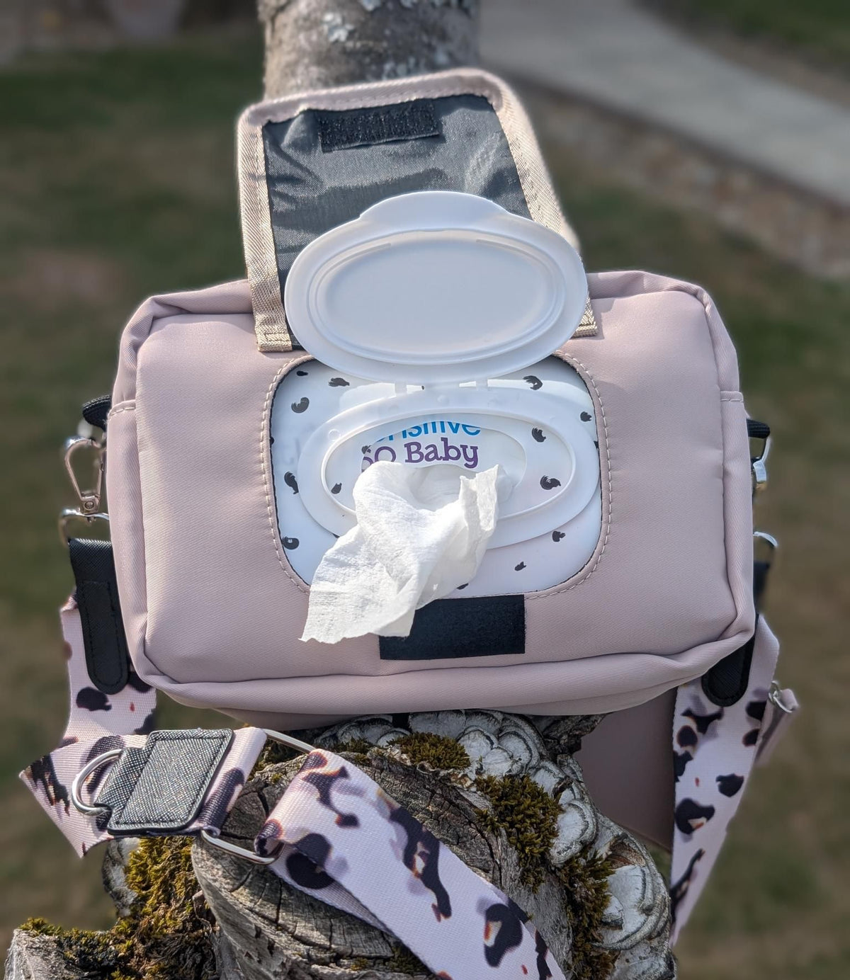 Pink diaper bag with a lid open, showing wipes inside, attached to a tree branch outdoors