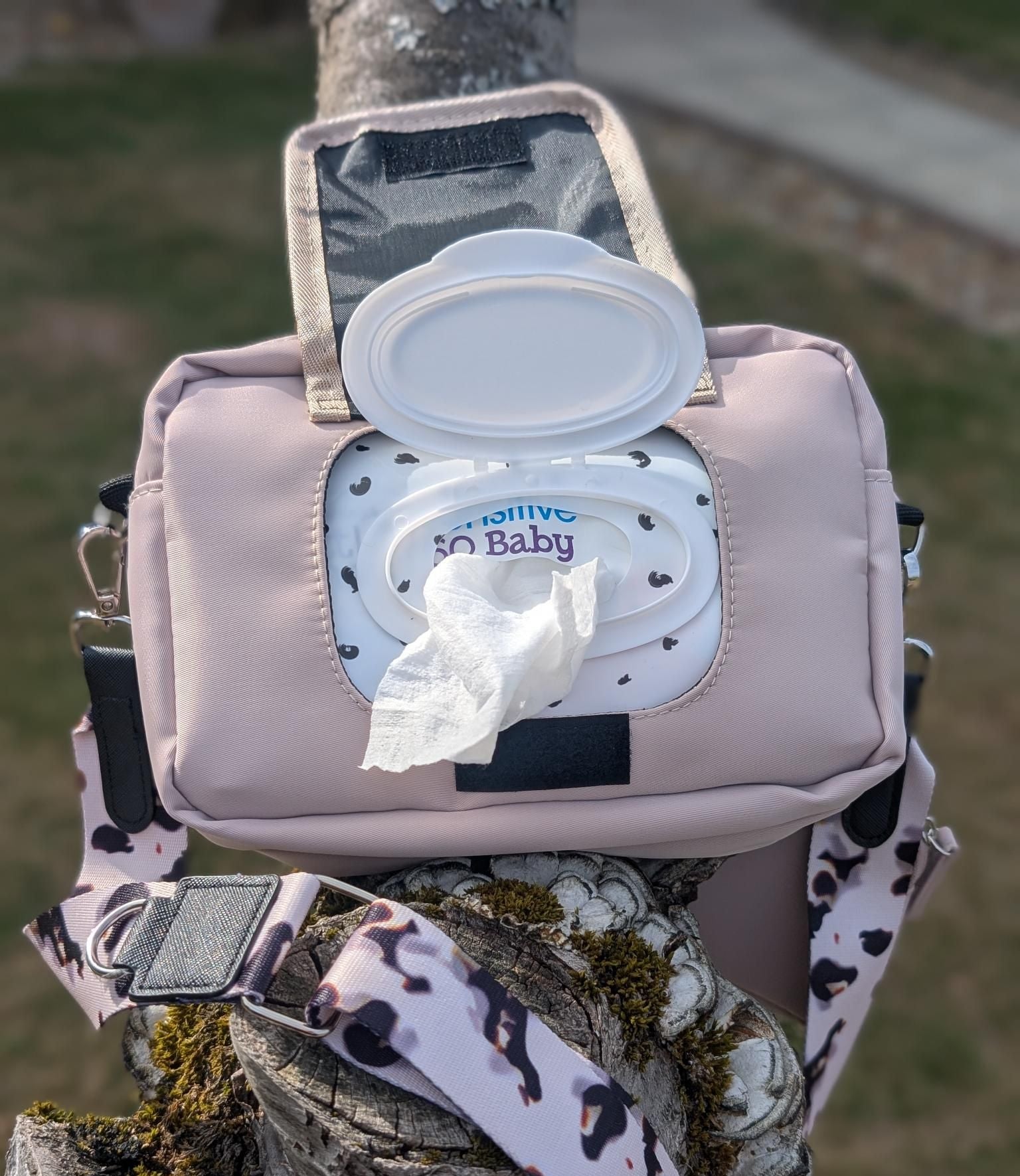 Pink diaper bag with a lid open, showing wipes inside, attached to a tree branch outdoors